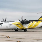 A imagem mostra um avião turboélice ATR da companhia aérea VOEPASS estacionado em um aeroporto urbano, com detalhes em amarelo e cinza sob um céu nublado.