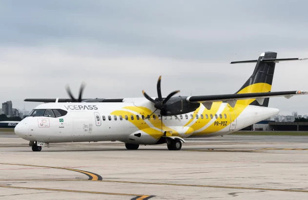A imagem mostra um avião turboélice ATR da companhia aérea VOEPASS estacionado em um aeroporto urbano, com detalhes em amarelo e cinza sob um céu nublado.