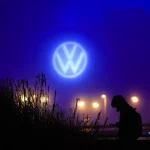 Uma pessoa segura um dispositivo sob o logotipo iluminado da Volkswagen em um cenário noturno com plantas e luzes ao fundo.