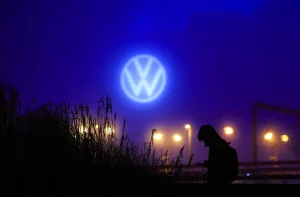 Uma pessoa segura um dispositivo sob o logotipo iluminado da Volkswagen em um cenário noturno com plantas e luzes ao fundo.