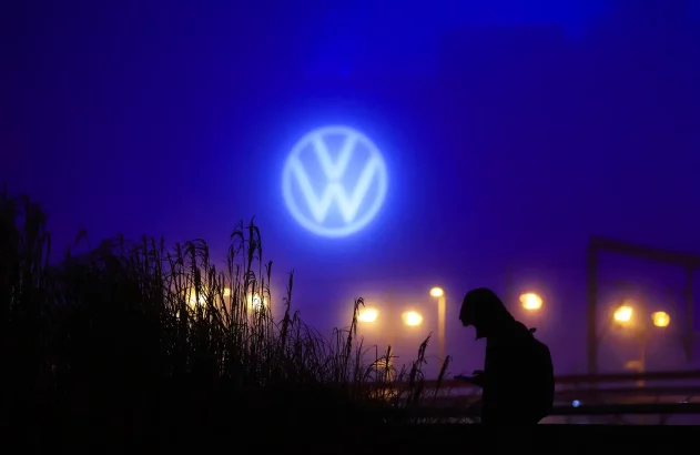 Uma pessoa segura um dispositivo sob o logotipo iluminado da Volkswagen em um cenário noturno com plantas e luzes ao fundo.