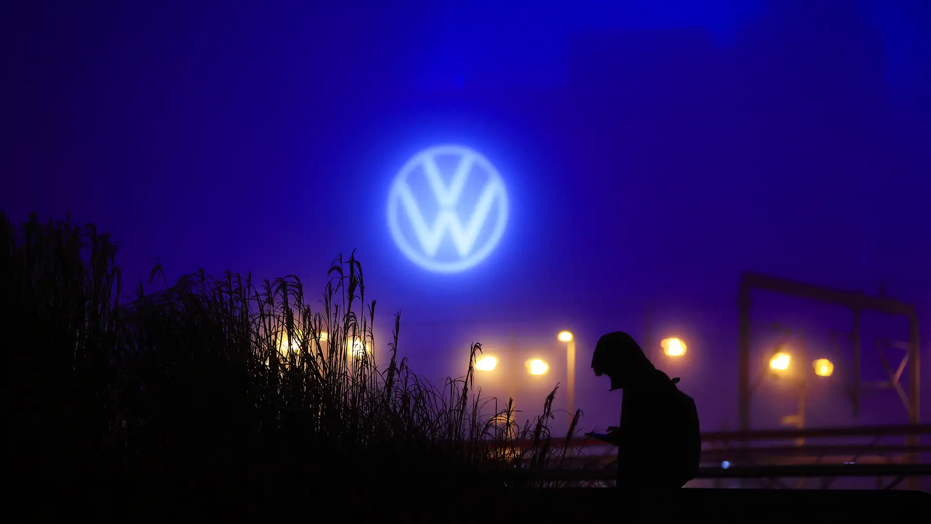 Uma pessoa segura um dispositivo sob o logotipo iluminado da Volkswagen em um cenário noturno com plantas e luzes ao fundo.