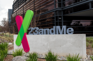 Placa da 23andMe com logotipo 'X' verde e rosa em frente a um edifício de escritórios moderno.