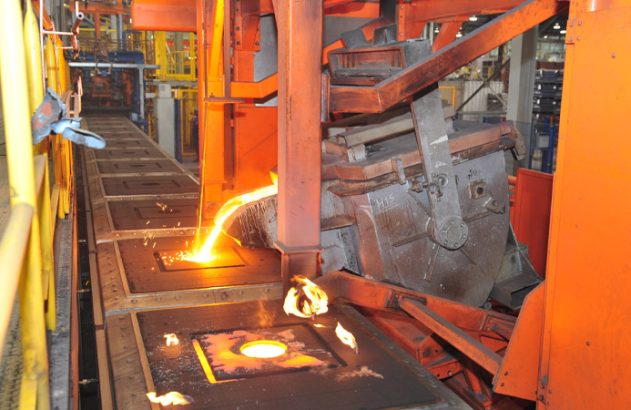 Metal fundido sendo despejado em moldes de uma esteira industrial, com brilho intenso e faíscas.