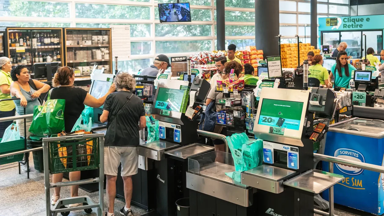 Interior de supermercado com caixas de autoatendimento e tradicionais, pessoas usando carrinhos de compras ou sacolas verdes, iluminação natural e prateleiras com lanches e bebidas; funcionários uniformizados em camisetas verdes.