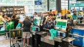 Interior de supermercado com caixas de autoatendimento e tradicionais, pessoas usando carrinhos de compras ou sacolas verdes, iluminação natural e prateleiras com lanches e bebidas; funcionários uniformizados em camisetas verdes.