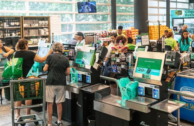 Interior de supermercado com caixas de autoatendimento e tradicionais, pessoas usando carrinhos de compras ou sacolas verdes, iluminação natural e prateleiras com lanches e bebidas; funcionários uniformizados em camisetas verdes.