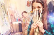 Menina sorridente com pincéis cobrindo parte do rosto em aula de arte. Outra criança pinta ao fundo.
