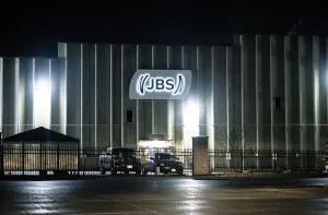 Fachada noturna do edifício industrial JBS com iluminação intensa e portão de segurança na entrada.