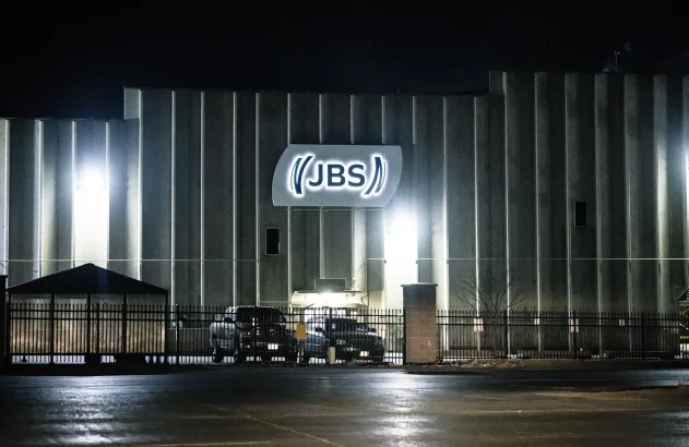 Fachada noturna do edifício industrial JBS com iluminação intensa e portão de segurança na entrada.