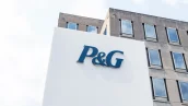 Imagem de um prédio corporativo moderno com a placa do logotipo azul da P&G em destaque, fachada minimalista em concreto cinza, janelas retangulares e céu parcialmente nublado.