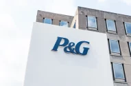 Imagem de um prédio corporativo moderno com a placa do logotipo azul da P&G em destaque, fachada minimalista em concreto cinza, janelas retangulares e céu parcialmente nublado.