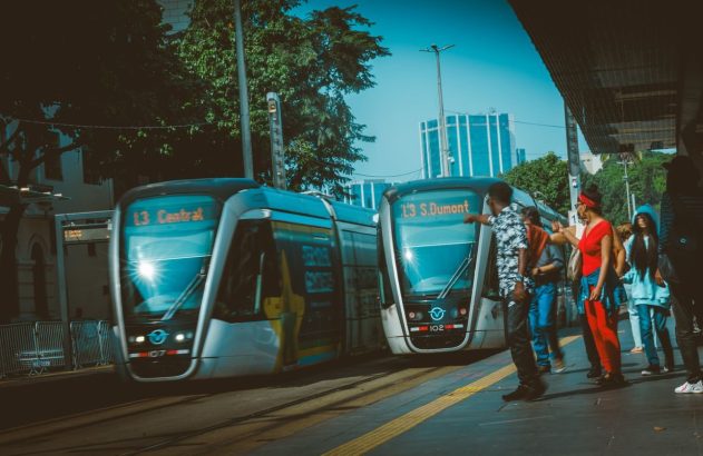 Dois VLTs em plataforma urbana. Um (13 Central) em movimento, outro (13 S.Dumont) parado, com pessoas.