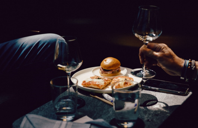 Duas pessoas em mesa escura com taças de vinho e prato de comida. Uma mão levanta a taça.