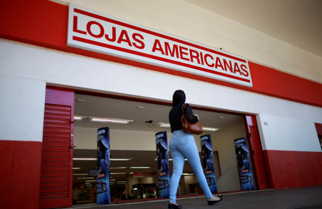 Mulher caminhando para a entrada da Lojas Americanas. Fachada vermelha e branca com letreiro da loja visível.