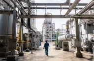 Imagem de uma instalação industrial complexa, semelhante a uma refinaria ou planta petroquímica, onde um trabalhador com roupa de proteção azul e capacete vermelho caminha entre tubulações de aço e uma torre de destilação ao fundo, evidenciando um ambiente de trabalho técnico, seguro e bem organizado.