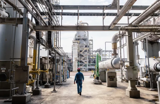 Imagem de uma instalação industrial complexa, semelhante a uma refinaria ou planta petroquímica, onde um trabalhador com roupa de proteção azul e capacete vermelho caminha entre tubulações de aço e uma torre de destilação ao fundo, evidenciando um ambiente de trabalho técnico, seguro e bem organizado.