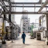 Imagem de uma instalação industrial complexa, semelhante a uma refinaria ou planta petroquímica, onde um trabalhador com roupa de proteção azul e capacete vermelho caminha entre tubulações de aço e uma torre de destilação ao fundo, evidenciando um ambiente de trabalho técnico, seguro e bem organizado.