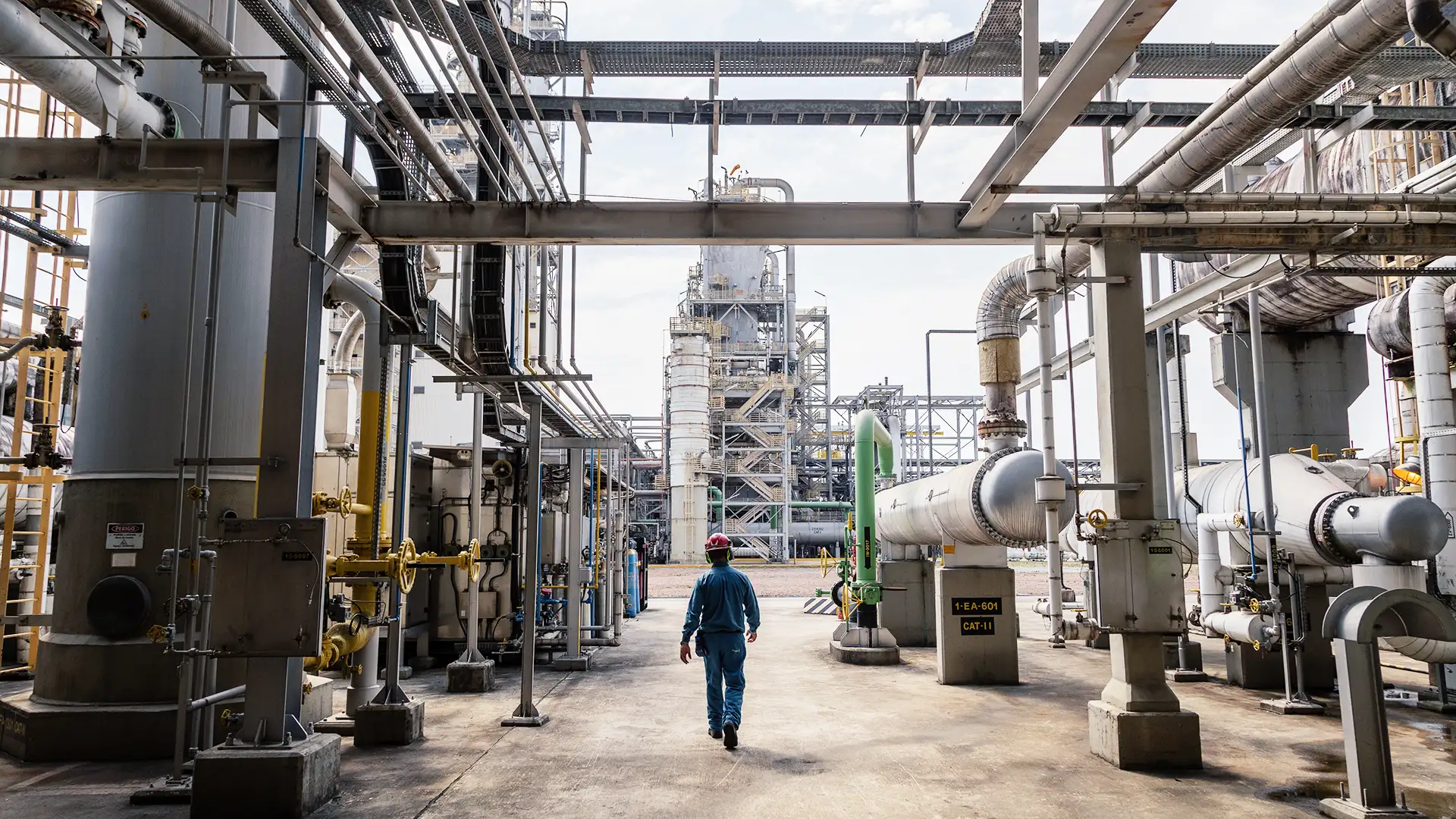 Imagem de uma instalação industrial complexa, semelhante a uma refinaria ou planta petroquímica, onde um trabalhador com roupa de proteção azul e capacete vermelho caminha entre tubulações de aço e uma torre de destilação ao fundo, evidenciando um ambiente de trabalho técnico, seguro e bem organizado.