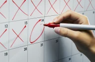 Imagem de uma mão segurando uma caneta vermelha enquanto circula a data 14 em um calendário de papel com fundo branco e números pretos. Datas 12 e 13 têm xis vermelhos.