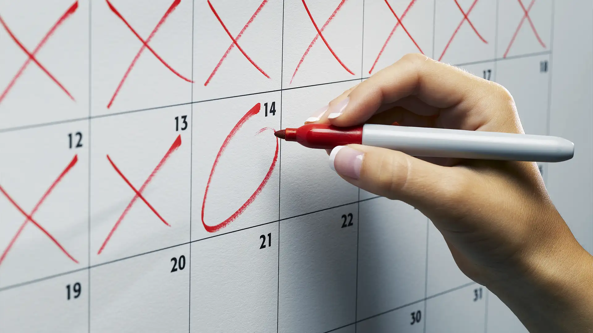 Imagem de uma mão segurando uma caneta vermelha enquanto circula a data 14 em um calendário de papel com fundo branco e números pretos. Datas 12 e 13 têm xis vermelhos.