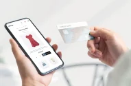Na imagem, uma pessoa segura um smartphone mostrando o site de compras "eshop" com um vestido vermelho e um cartão de crédito na outra mão, pronta para finalizar a compra online.