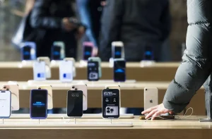Exposição de iPhones em loja, com cliente tocando um aparelho. Telas exibem conteúdo como 'Apple Intelligence'.