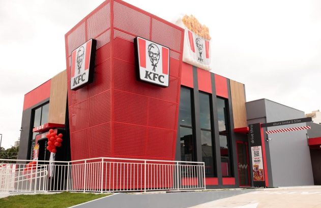 Exterior de um restaurante KFC moderno, com fachada vermelha e cinza, e balde gigante de frango no topo.