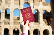 Imagem de uma mão segurando um passaporte italiano com letras douradas, destacando o emblema da República Italiana, com o Coliseu de Roma ao fundo sob um céu azul sem nuvens.