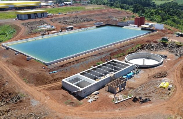 Vista aérea de estação de tratamento de água em construção, com bacia azul, tanque e estruturas de concreto.