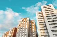 Três edifícios altos com fachada nas cores marrom, creme e cinza sob um céu azul com nuvens. Janelas alinhadas e linhas retas predominam na arquitetura urbana contemporânea.