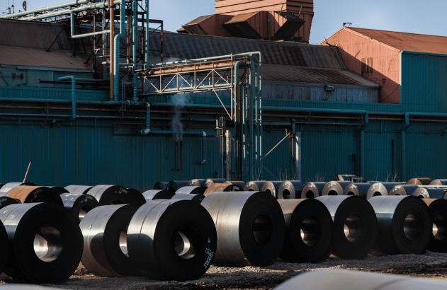 Pátio industrial com bobinas de aço. Fábrica ao fundo, com tubulações e vapor saindo.