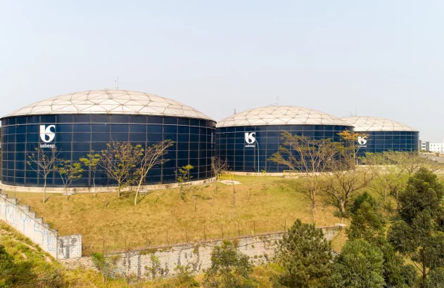 Três grandes reservatórios cilíndricos azuis com o logotipo da Sabesp estão cercados por vegetação e um muro grafitado, próximos a uma área urbana.