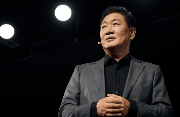 Homem de terno escuro em palco, com microfone e mãos unidas, olhando atento.