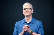 Tim Cook de polo azul, óculos e Apple Watch, com as mãos juntas, olhando para o lado.