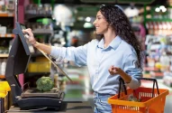 Na imagem, uma mulher com cabelo cacheado solto, vestindo camisa azul clara e jeans, está em um supermercado pesando um brócolis em uma balança e interagindo com um monitor ao lado, enquanto segura uma cesta de compras laranja.