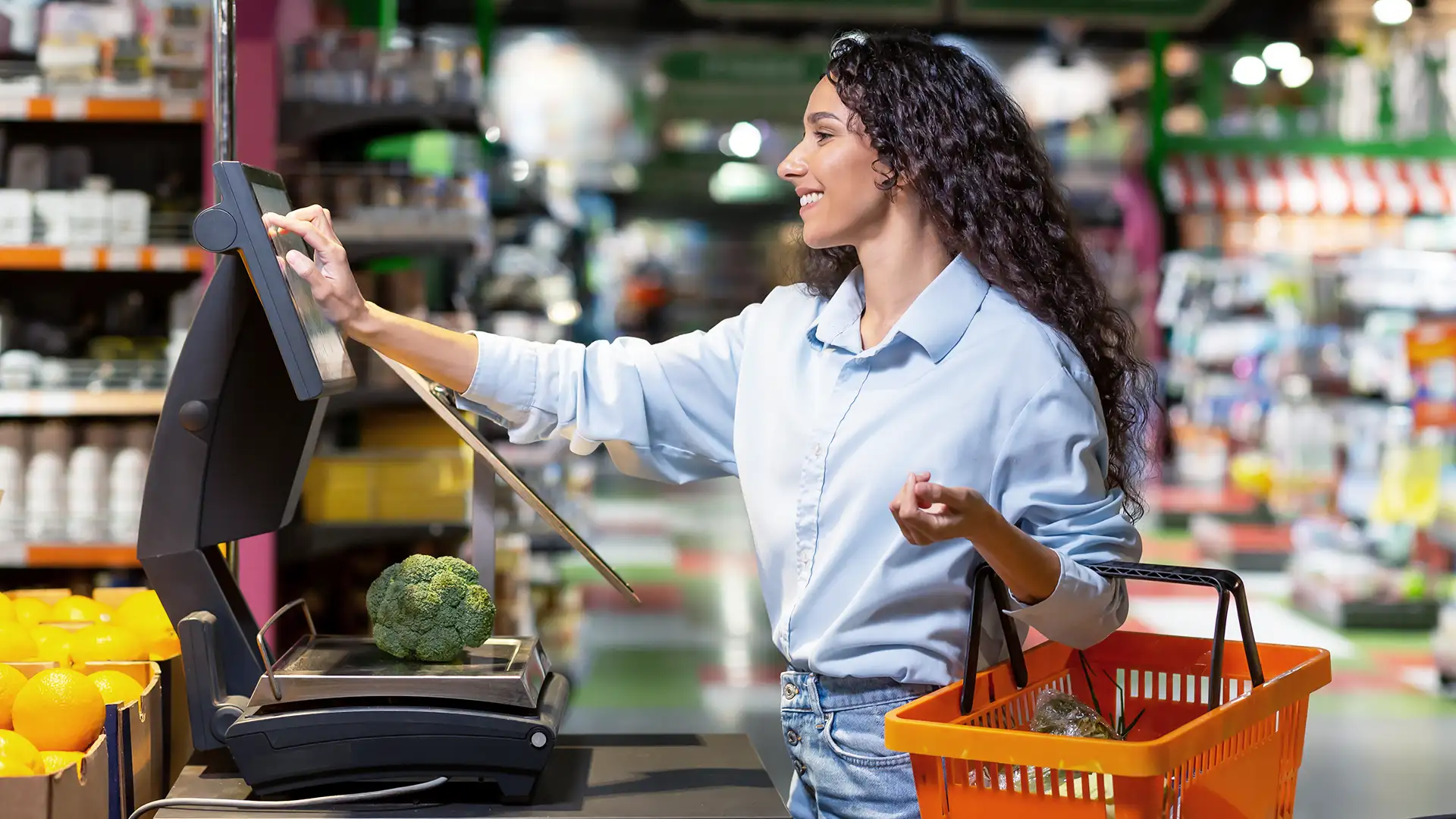 Na imagem, uma mulher com cabelo cacheado solto, vestindo camisa azul clara e jeans, está em um supermercado pesando um brócolis em uma balança e interagindo com um monitor ao lado, enquanto segura uma cesta de compras laranja.