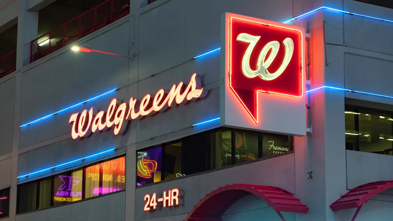 Fachada de loja Walgreens em neon, com letreiro vermelho cursivo e logotipo vermelho e branco, detalhes modernos em neon azul e inscrição "24-HR".