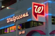 Fachada de loja Walgreens em neon, com letreiro vermelho cursivo e logotipo vermelho e branco, detalhes modernos em neon azul e inscrição "24-HR".