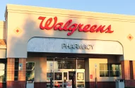 Fachada da loja Walgreens com logo vermelho e letreiro "PHARMACY", em tons claros e ladrilhos, portas automáticas, sinal de vacinas contra gripe e estacionamento acessível; foto ao entardecer.