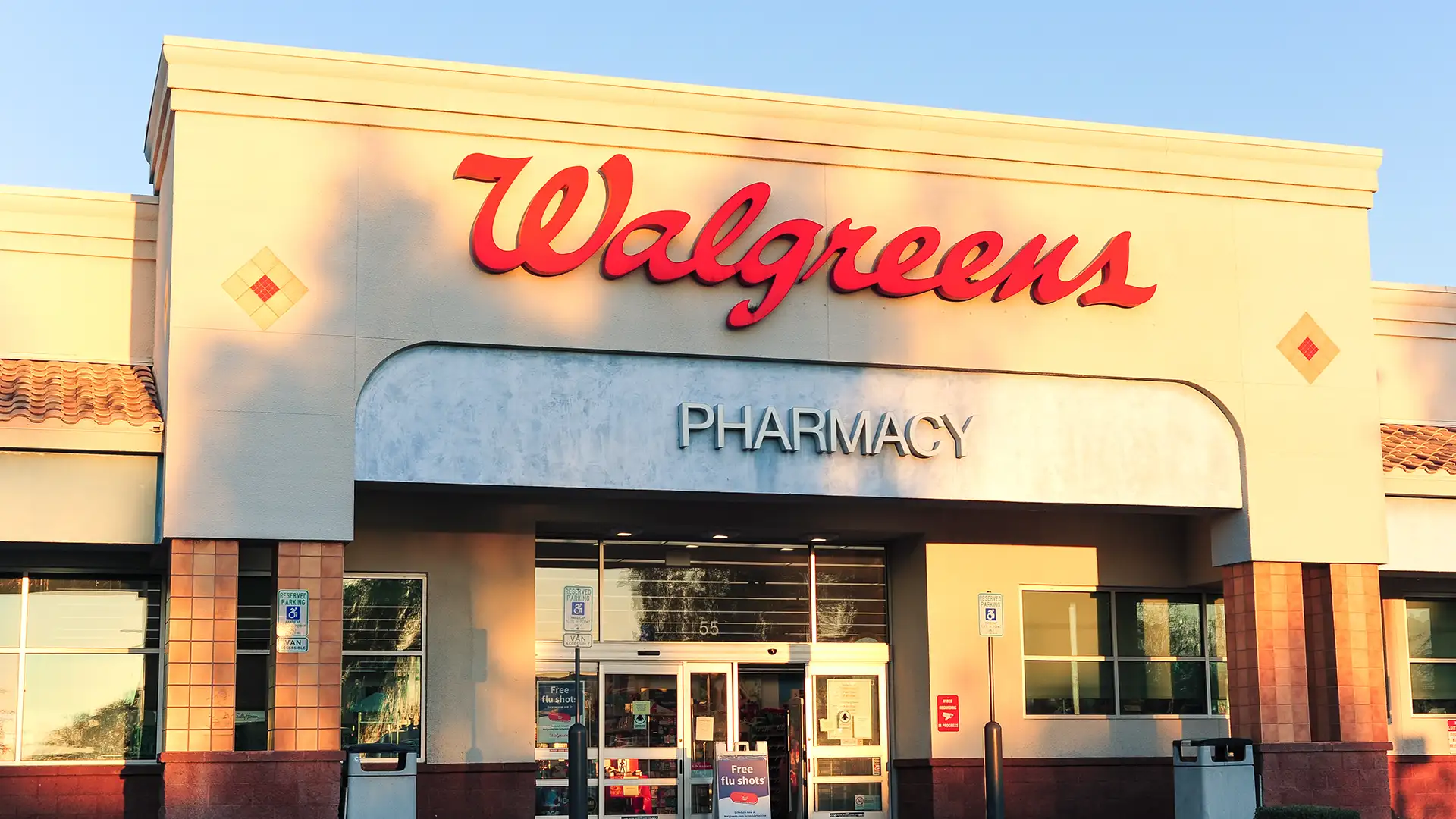 Fachada da loja Walgreens com logo vermelho e letreiro "PHARMACY", em tons claros e ladrilhos, portas automáticas, sinal de vacinas contra gripe e estacionamento acessível; foto ao entardecer.