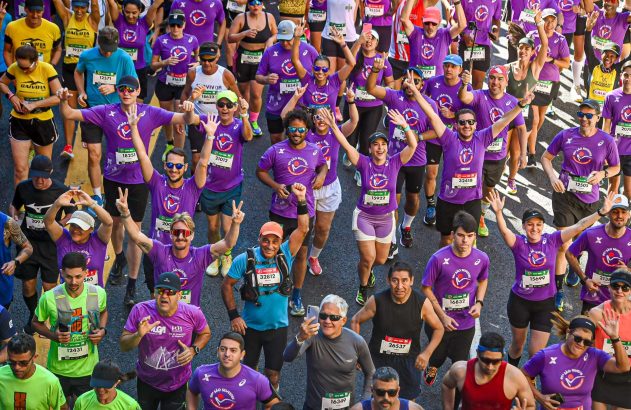 Multidão de corredores com camisetas roxas sorrindo e acenando em uma corrida de rua, a 99ª São Silvestre.