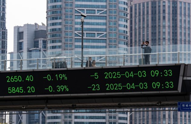 Painel LED exibe cotações da bolsa. Pessoas em passarela, uma tira foto. Prédios modernos da cidade ao fundo.