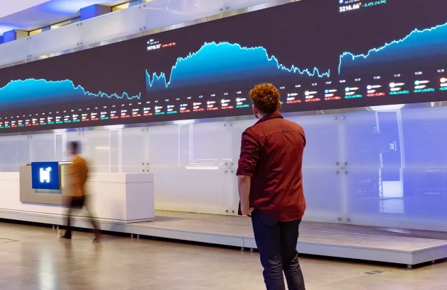 Homem de costas observa tela gigante com gráficos de mercado financeiro. Uma pessoa borrada transita em primeiro plano.