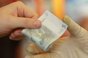 Uma mão sem luva entrega uma nota de 5 euros para uma mão enluvada.