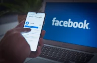 Uma pessoa interage com o aplicativo do Facebook em um smartphone, com um laptop exibindo o logotipo do Facebook ao fundo.