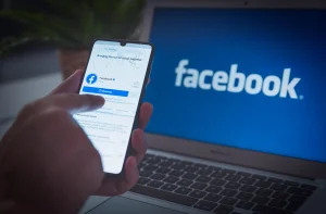 Uma pessoa interage com o aplicativo do Facebook em um smartphone, com um laptop exibindo o logotipo do Facebook ao fundo.