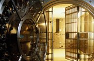 Grande porta de cofre de banco redonda e aberta, com engrenagens visíveis, e grade de segurança revelando o interior.