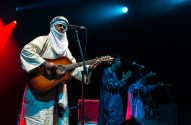 Homem com turbante e túnica clara toca violão em palco escuro com luzes vermelhas e azuis. Outros músicos ao fundo.