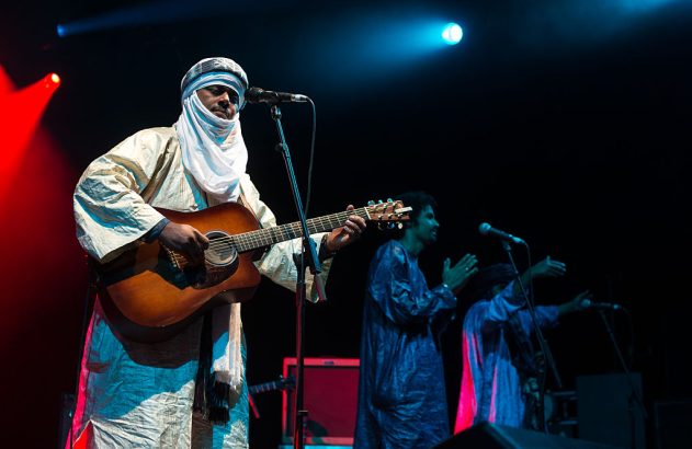 Homem com turbante e túnica clara toca violão em palco escuro com luzes vermelhas e azuis. Outros músicos ao fundo.